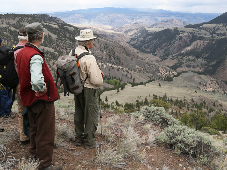A group of field naturalists stand near an edge overlooking a river river valley far below surrounding by a grassy canyon with sparse bushes and trees.