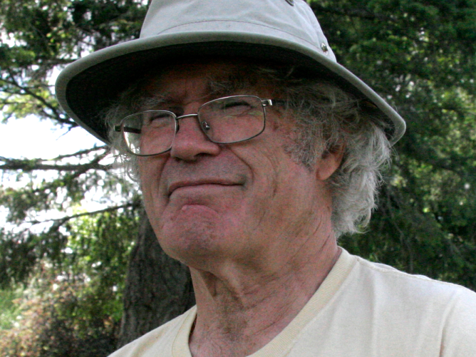 Close up of Fred McMechan outdoors with a hat and glasses, backdropped by a dark green tree.