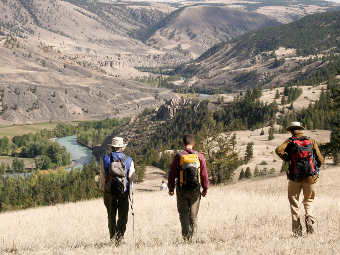 Three field naturalists descend a grassy slope towards Farwell Canyon's turquoise river, which meanders its way through the canyon and grasslands. Along the edge are lush green trees and bushes.